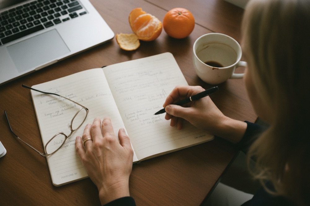 Looking down at a desk: an open notebook, reading glasses, a cup of coffee and tangerines, with a hand writing