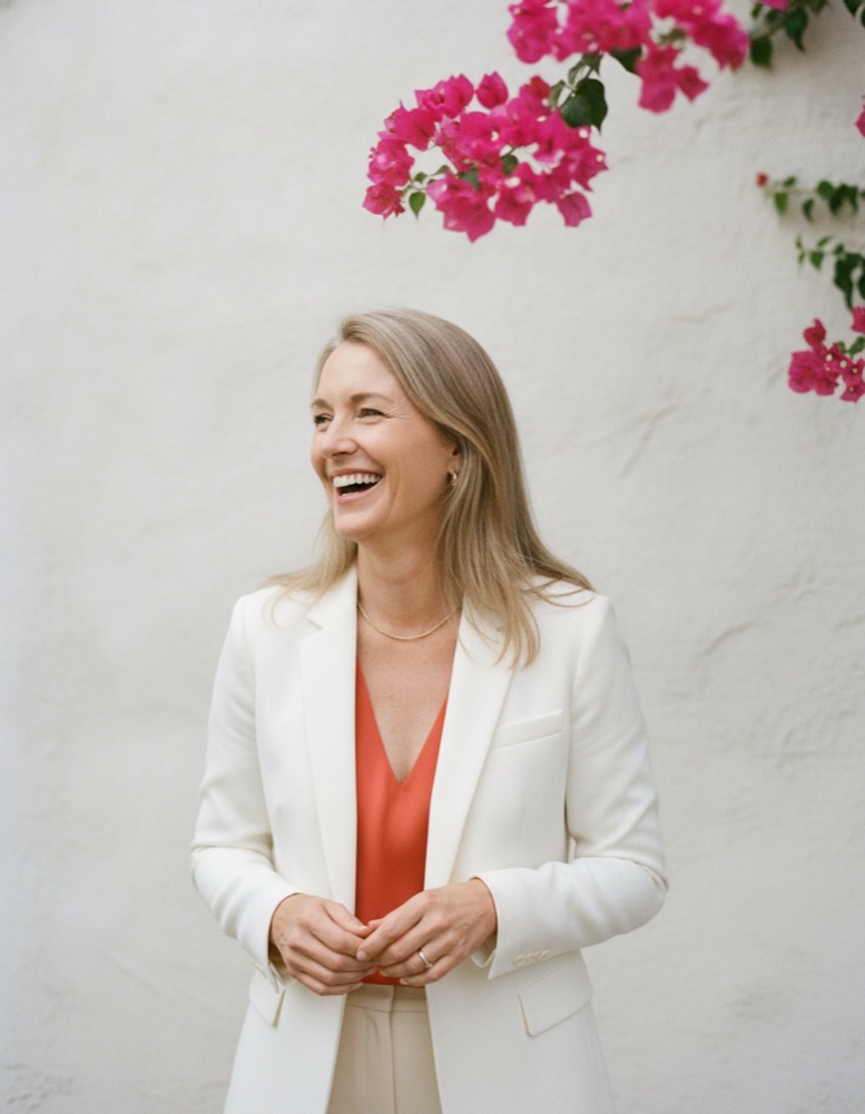 Claire laughing against a white stucco wall with pink bougainvillea, in a white blazer and tangerine top